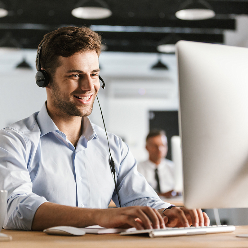 Ein Mann aus dem Notruf Service Center des Unternehmens ÖWD Security & Services mit Headset arbeitet lächelnd an einem Computer in einem Büro.