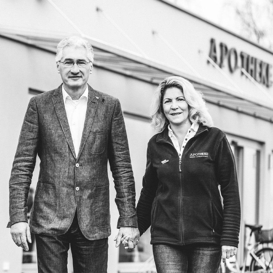 Ein Mann und eine Frau stehen vor einem Gebäude mit dem Schriftzug "Apotheke" und halten Hände.