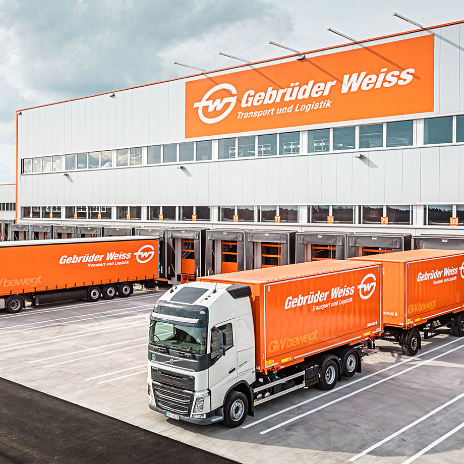 Ein Lastwagen von Gebrüder Weiss steht vor einer Laderampe eines Logistikzentrums. Auf dem Gebäude und den Anhängern ist das Firmenlogo zu sehen.