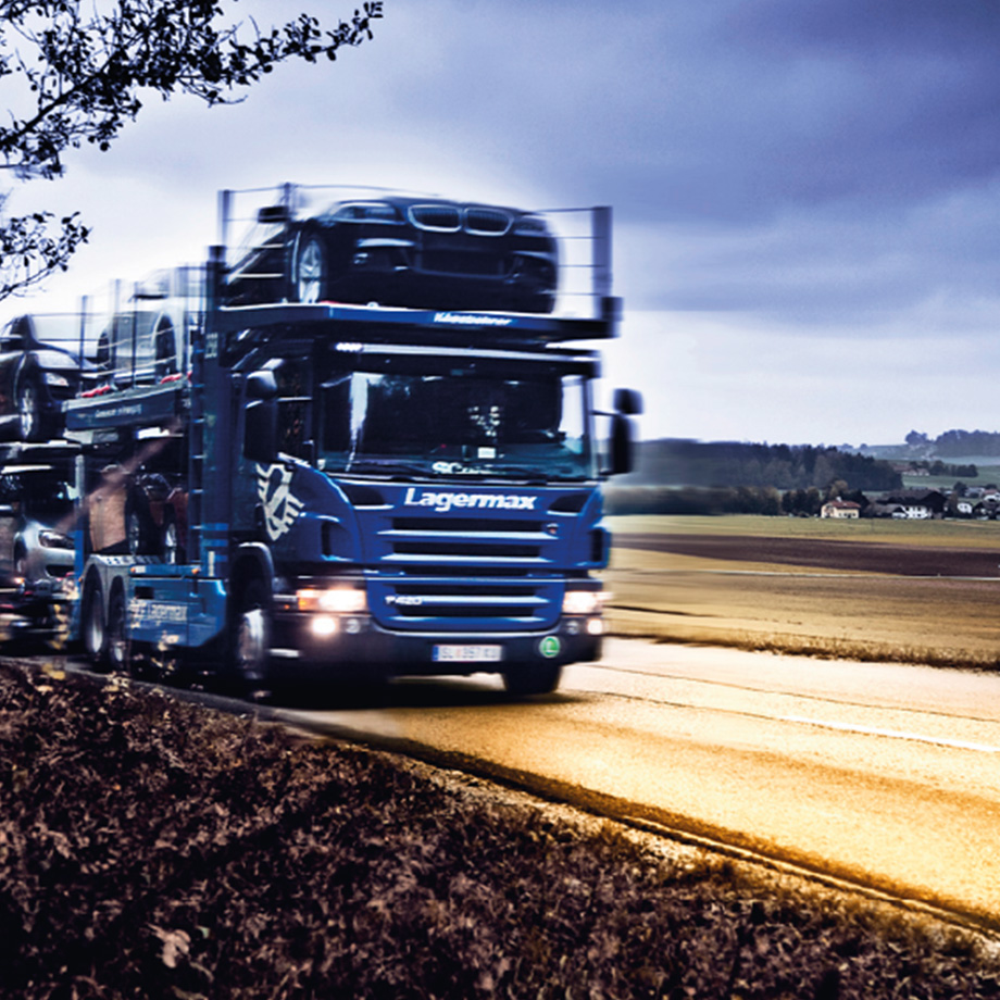 Ein Autotransporter von Lagermax fährt auf einer Landstraße, mit Autos auf dem Anhänger gegen einen bewölkten Himmel.