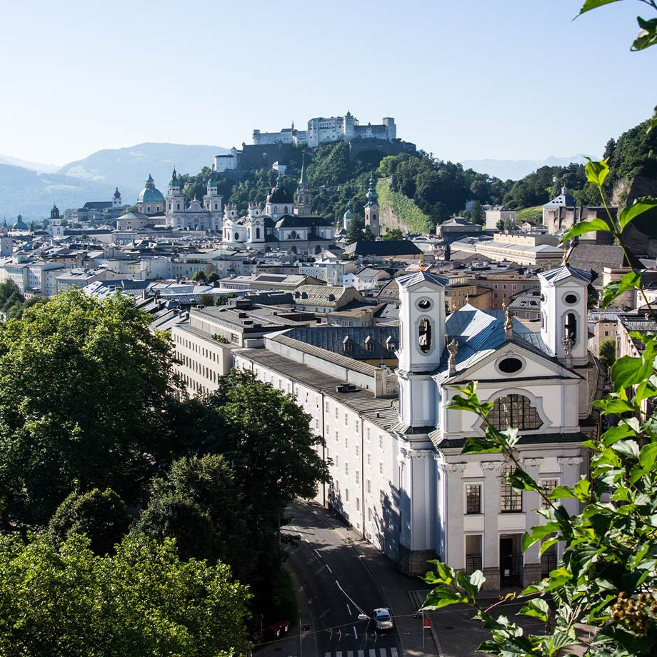 Panorama von Salzburg mit der Festung Hohensalzburg im Hintergrund und grünen Bäumen im Vordergrund.