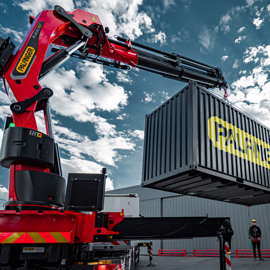 Ein roter Kran der Firma Palfinger hebt einen großen Container in einem Industriegelände.